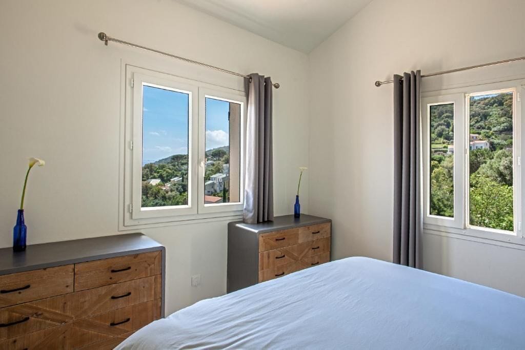 Quarto com cama, cómoda de madeira e duas janelas com cortinas.