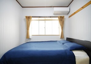 A room with a bed, a window with curtains, an air conditioning unit, and a wooden beam on the wall.