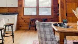 Interior acogedor de una cabaña con un gran ventanal central, paredes de madera, un comedor y una cocina con armarios blancos.