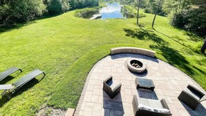 Un área de estar circular al aire libre con un brasero, rodeada por un patio empedrado, frente a un césped y una masa de agua.
