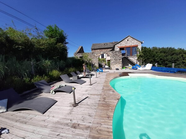 A wooden deck with lounge chairs, a swimming pool, and a stone building with a red roof.