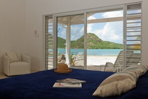 Une chambre à coucher avec un lit, une chaise blanche et une vue sur la plage et les montagnes, visible à travers de grandes portes vitrées.