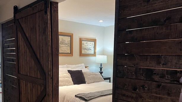 A bedroom with a large wooden sliding door, a bed with white bedding, a bedside table with a lamp, and two framed pictures on the wall. - Cozy Cabin (Red River)