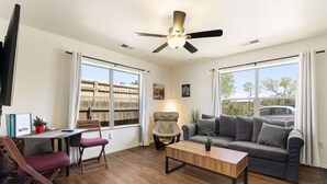 Une pièce avec un plancher en bois, un ventilateur de plafond, un canapé gris, une table basse en bois, un bureau avec une chaise et de grandes fenêtres avec des rideaux.
