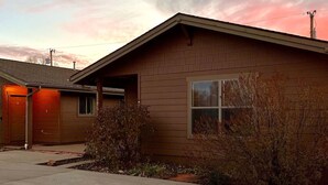 Une maison de plain-pied à l’extérieur brun, avec une porte de garage et un petit jardin devant, agrémenté de quelques arbustes.