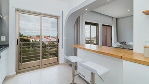 Une cuisine moderne avec un plan de travail en bois, deux tabourets de bar blancs et une porte-fenêtre coulissante donnant sur un balcon avec vue sur la ville.