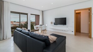 Ein modernes Wohnzimmer mit einem schwarzen Ledersofa, einem an der Wand montierten Flachbildfernseher und einem Essbereich mit einem weißen Tisch und Stühlen.
