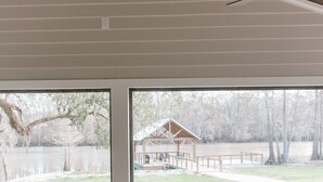 A covered deck with a view of a lake and a wooden structure in the distance.