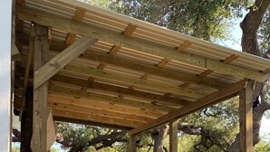 Et overdækket udendørs opholdsområde med et træpergola, en polstret sofa og en fletstol.