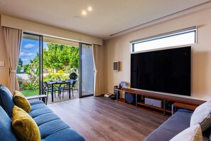 A living room with a large TV, a sofa, a coffee table, and a sliding glass door leading to a garden.