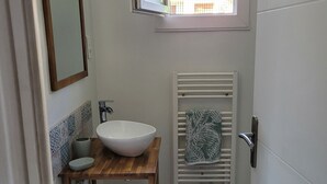 A bathroom with a wooden vanity, a white sink, a mirror, a towel rack, and a window with a view of buildings. - T2 near historic center (Manosque)