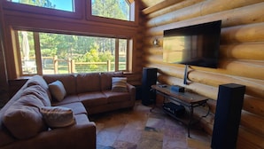 A cozy log cabin interior with a sofa, a flat-screen TV mounted on the wall, and a large window offering a view of the outdoors. - Spacious cabin in Angel Fire area - 6 miles to ski slopes and recreation area (Angel Fire)