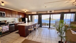 Une cuisine moderne dotée d’un îlot central, d’un coin repas avec des tabourets de bar et d’une grande porte-fenêtre coulissante donnant sur un balcon avec vue sur la mer.