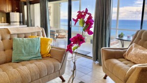 Un salon comprenant un canapé, des fauteuils et une table basse. De grandes fenêtres offrent une vue sur l’océan.