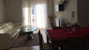 Ett vardagsrum med en beige soffa, ett soffbord med glasskiva, ett matbord täckt med en röd duk och en röd stol.
