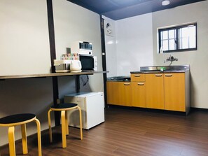 A compact kitchen with a microwave, small refrigerator, and wooden cabinets. There are two stools and a window providing natural light.