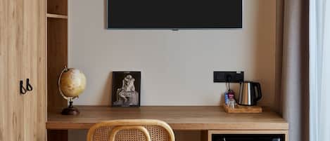 A compact room with a wooden desk, a black TV mounted on the wall, a globe, a framed picture, a small coffee maker, and a compact refrigerator.