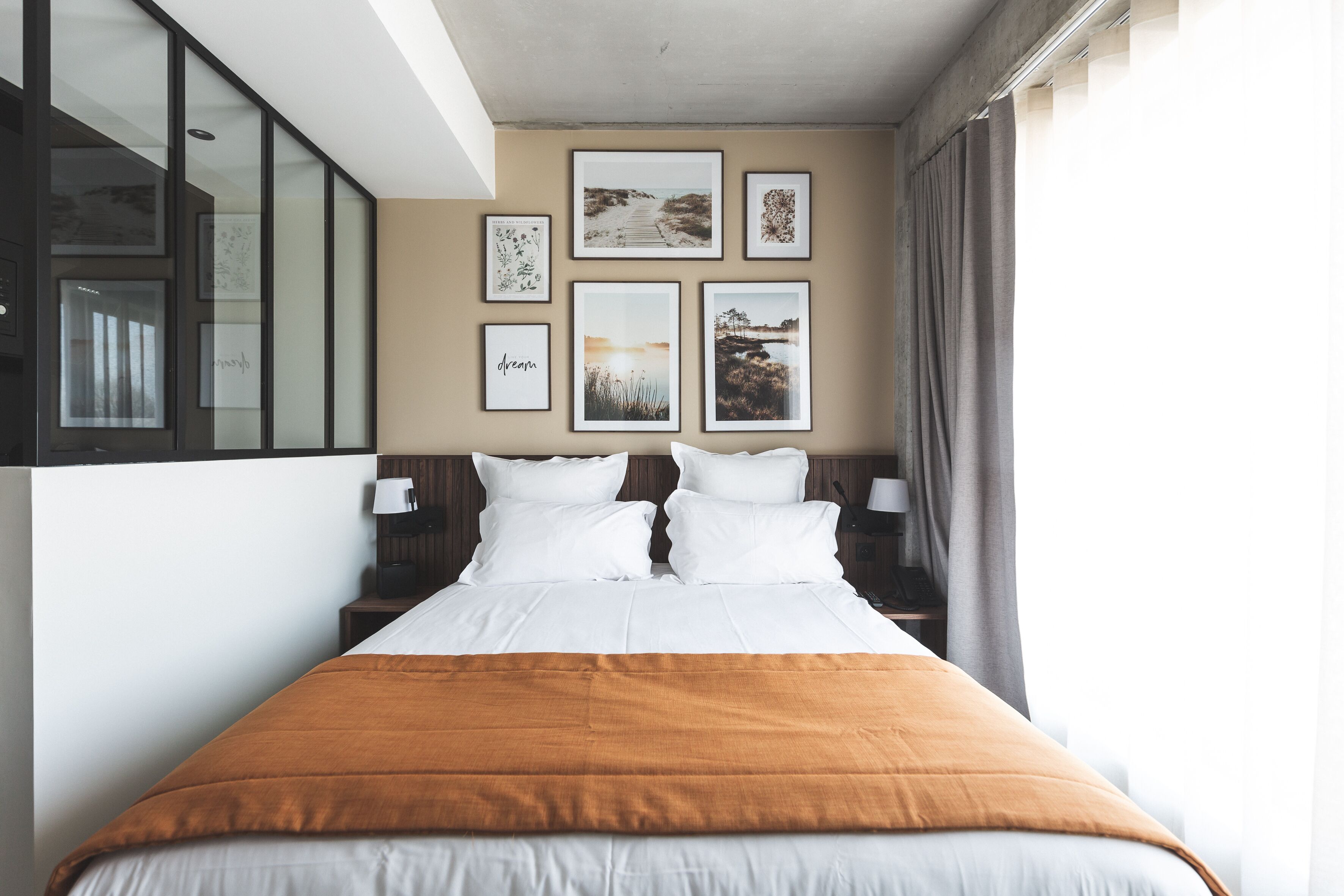 A hotel room with a bed, two bedside tables, a window with curtains, and a wall with framed pictures.