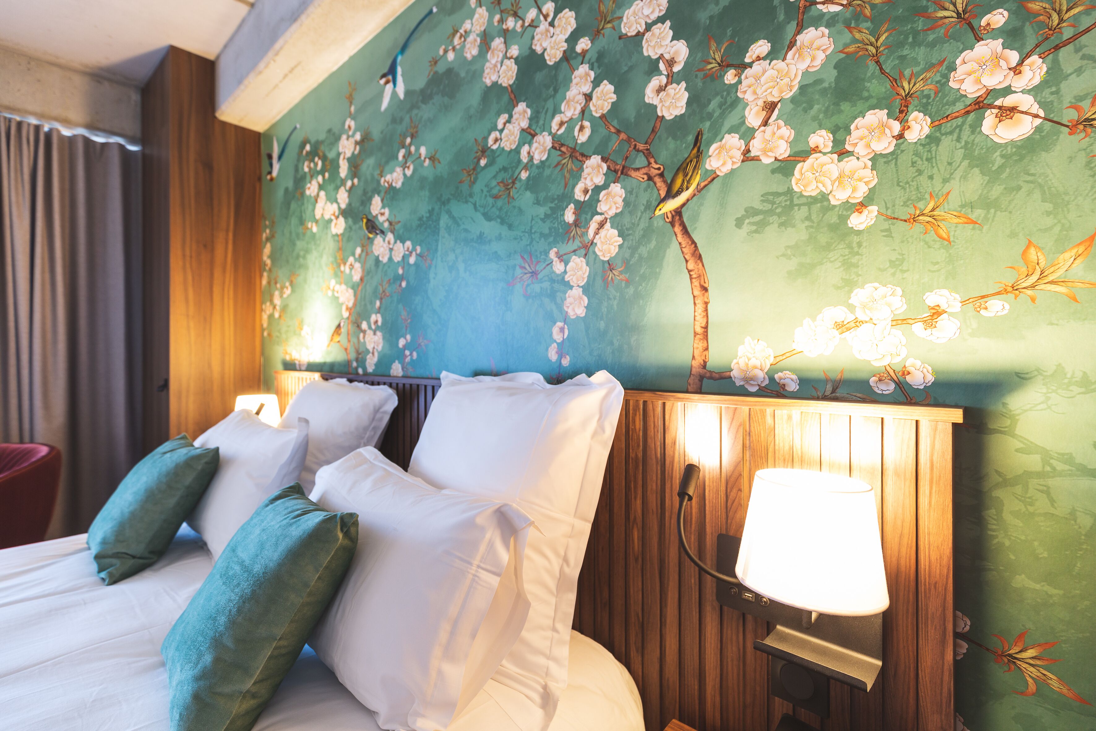 Une chambre d’hôtel avec un lit, des oreillers, une table de chevet, une lampe et une fresque murale représentant des fleurs de cerisier.