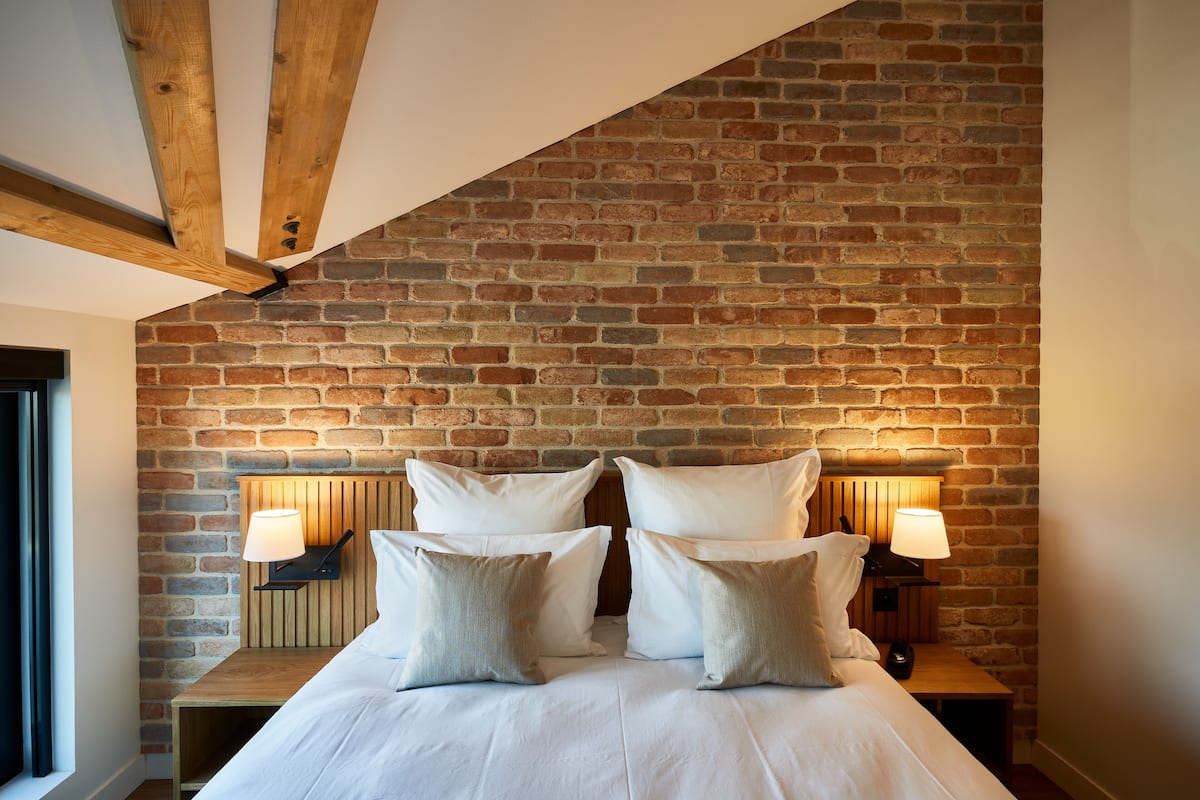 A bedroom with a brick wall, wooden beams, a bed with white linens, and two bedside lamps.