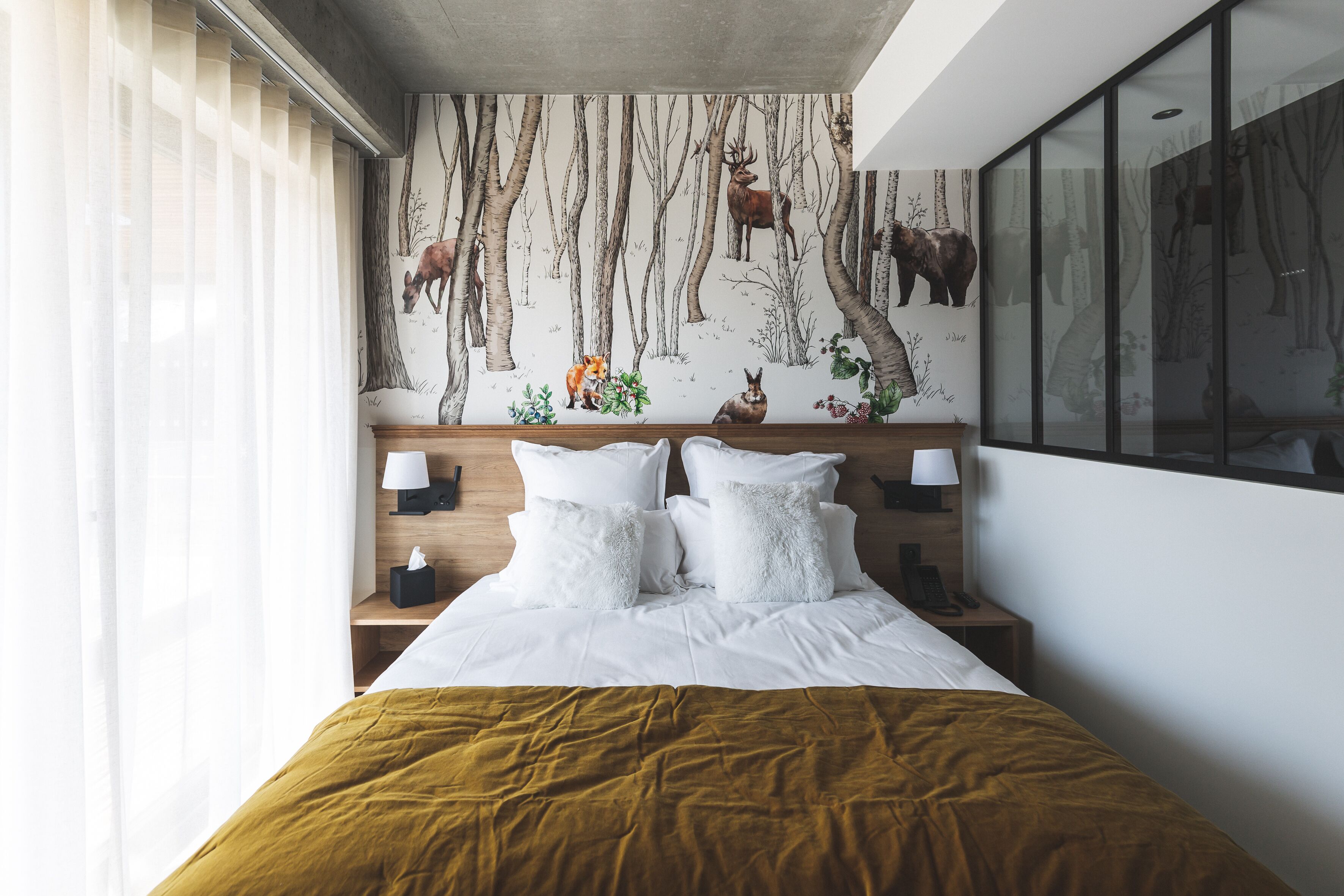 Ein modernes Hotelzimmer mit einem großen Bett, weißer Bettwäsche, einer senffarbenen Tagesdecke, einem Wandgemälde mit Waldmotiv und einem großen Fenster mit Jalousien.