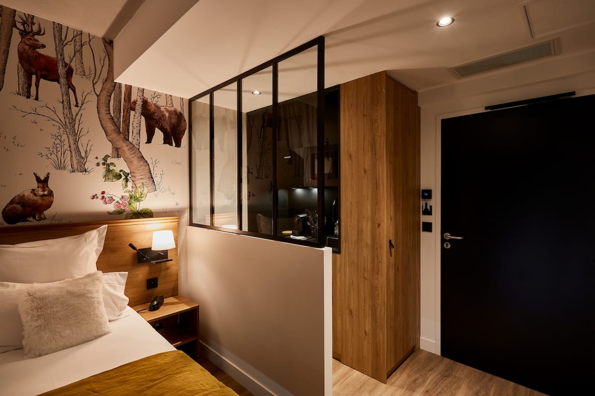 A hotel room with a bed, a nightstand, a lamp, a wooden wardrobe, a dark door, and a wall mural of deer and trees.