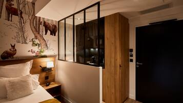 A hotel room with a bed, a nightstand, a lamp, a wooden wardrobe, a dark door, and a wall mural of deer and trees.