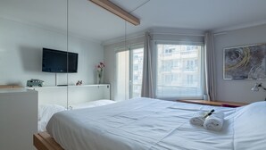 Una habitación de hotel moderna con una cama grande, un televisor de pantalla plana, una mesita de noche y un ventanal con persianas.