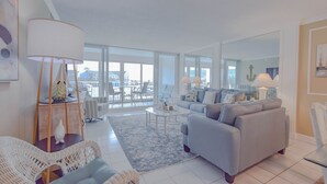 Una sala moderna con un sofá gris, una mesa de centro blanca y sillones de mimbre. Amplias ventanas ofrecen vistas al exterior.