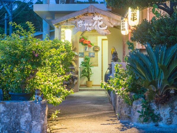 Uma entrada tradicional japonesa com portão de madeira, plantas em vasos e um caminho de pedras até a porta.