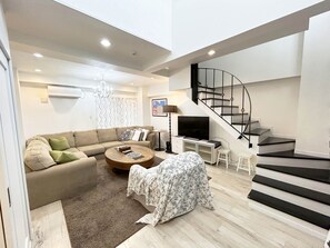 Uma sala de estar moderna com um sofá, mesa de centro e um banquinho com tampo. Uma escada com corrimão preto leva a um andar superior.