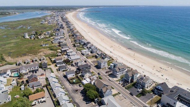 Vue aérienne d’une zone résidentielle côtière, avec des maisons le long de la plage et une route qui longe le rivage.