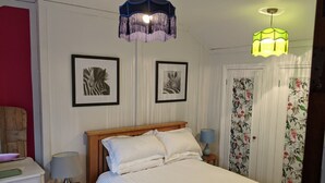 A neatly arranged bedroom with a bed, two framed pictures, a nightstand, a chair, a lamp, and a door with floral curtains. - Lovely Stables mew's house in hidden town location (Royal Tunbridge Wells)