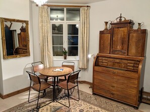 A dining area with a round wooden table and four chairs, a wooden cabinet with drawers, a mirror with an ornate frame, and a window with curtains.