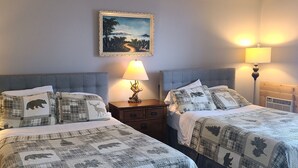 Habitación de hotel con dos camas, cada una con cabecero gris, una mesita de noche entre ellas y dos lámparas de noche. Un cuadro enmarcado cuelga en la pared sobre las camas.