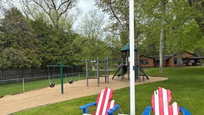 Un parque infantil con columpios, un tobogán y una estructura para escalar. Hay dos sillas Adirondack y una mesita.