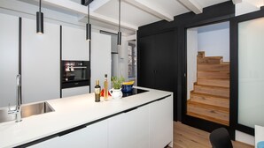 Une cuisine moderne dotée d’un îlot central, de luminaires suspendus noirs et d’un escalier en bois menant à un balcon.