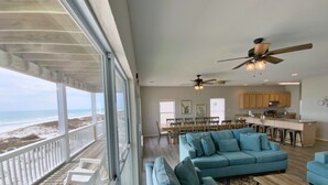 Een ruime woonkamer met een grote blauwe bank, een houten salontafel en uitzicht op het strand door grote ramen.
