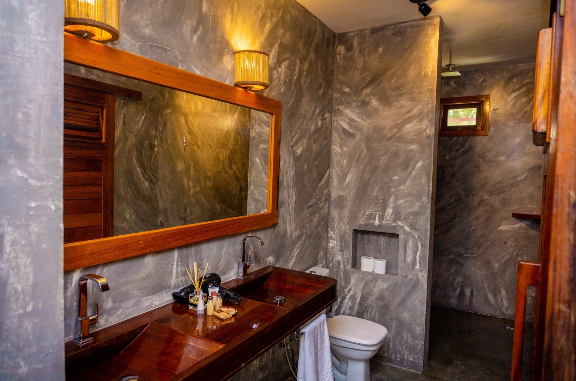 Un baño con pared de mármol, tocador de madera, un espejo grande, lavamanos, inodoro y zona de ducha.