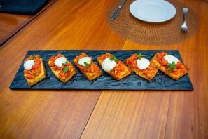 Plato rectangular con seis bruschettas cubiertas de tomates y mozzarella, acompañado de un plato blanco y un tenedor sobre una mesa de madera.