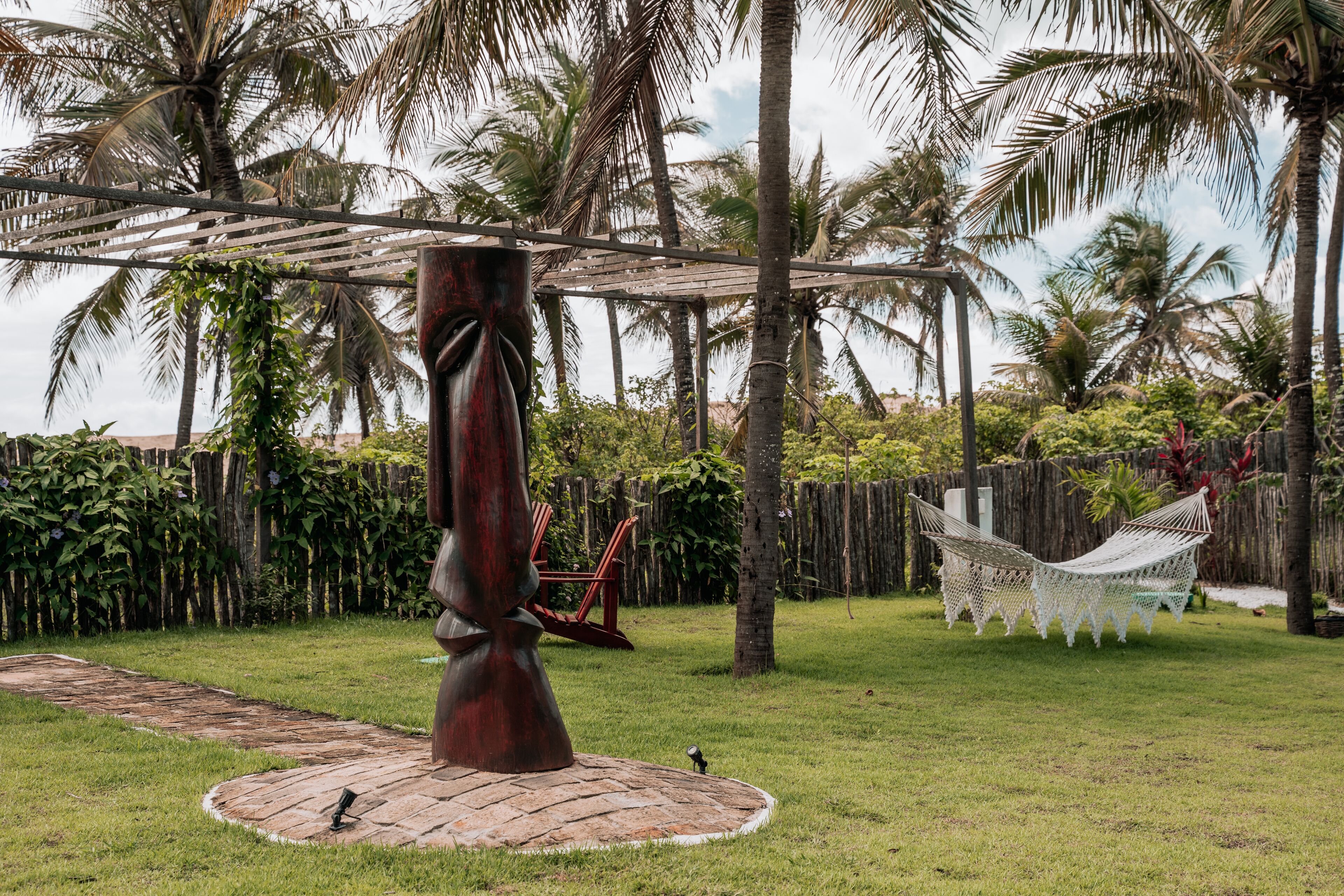 A hammock under a wooden pergola in a garden with palm trees and a wooden fence.