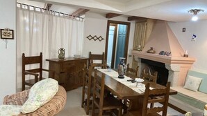 A dining area with a wooden table, chairs, and a wicker chair. There is a fireplace with decorative items on the mantel.