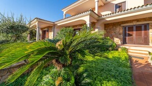 Une villa de style méditerranéen, dotée d’une façade en pierre, d’un porche abrité et d’une végétation luxuriante au premier plan.