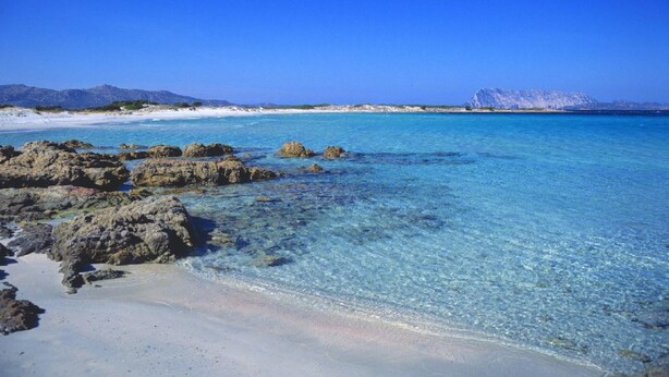 Une plage bordée d’une eau turquoise et limpide, de formations rocheuses et dominée par des montagnes lointaines.