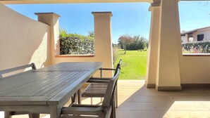Un patio exterior con una mesa y sillas de madera, vista a un césped verde y un edificio al fondo.