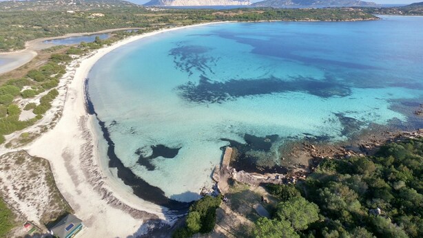 Vista aérea de una zona costera con una playa de arena, aguas azul cristalino y montañas al fondo.