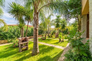 Ein gepflegter Garten mit Palmen, einer Steinsitzbank und einem Weg, der zu einem Gebäude mit einer Steinfassade führt.