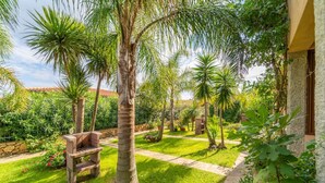 Ein gepflegter Garten mit Palmen, einer Steinsitzbank und einem Weg, der zu einem Gebäude mit einer Steinfassade führt.