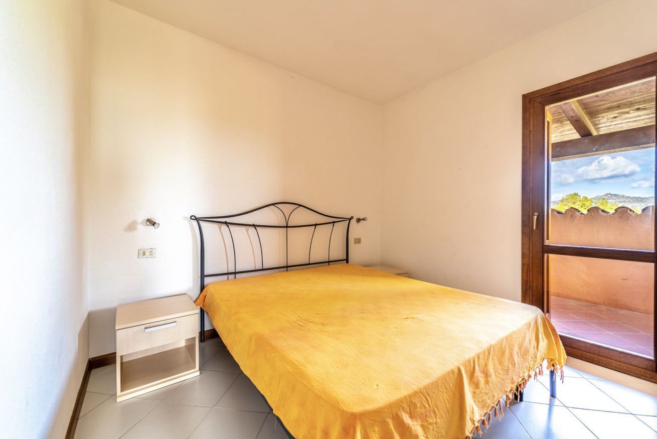 Un lit simple avec un cadre en métal et une couvre-lit jaune, dans une pièce aux murs blancs et au sol carrelé.