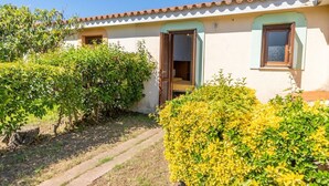 Uma casa com telhado de telhas, uma porta e uma janela. Há uma passagem que leva à casa e um arbusto em primeiro plano.